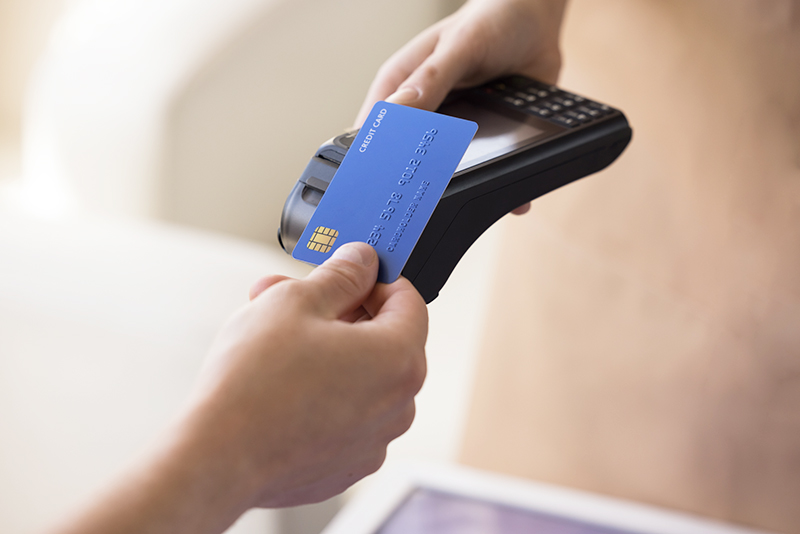 Hand of bank cardholder paying bill in cafe, applying blue credit card with chip at wireless payment terminal held by waitress, using electronic transaction banking technology. Clopped shot, close up